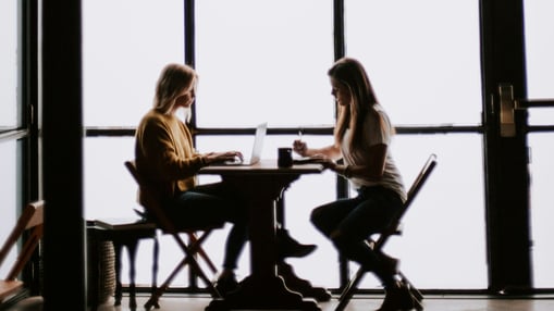 Duas mulheres sentadas em uma mesa, uma de frente para a outra. Uma delas está com um notebook e a outra com uma caneta e papel.