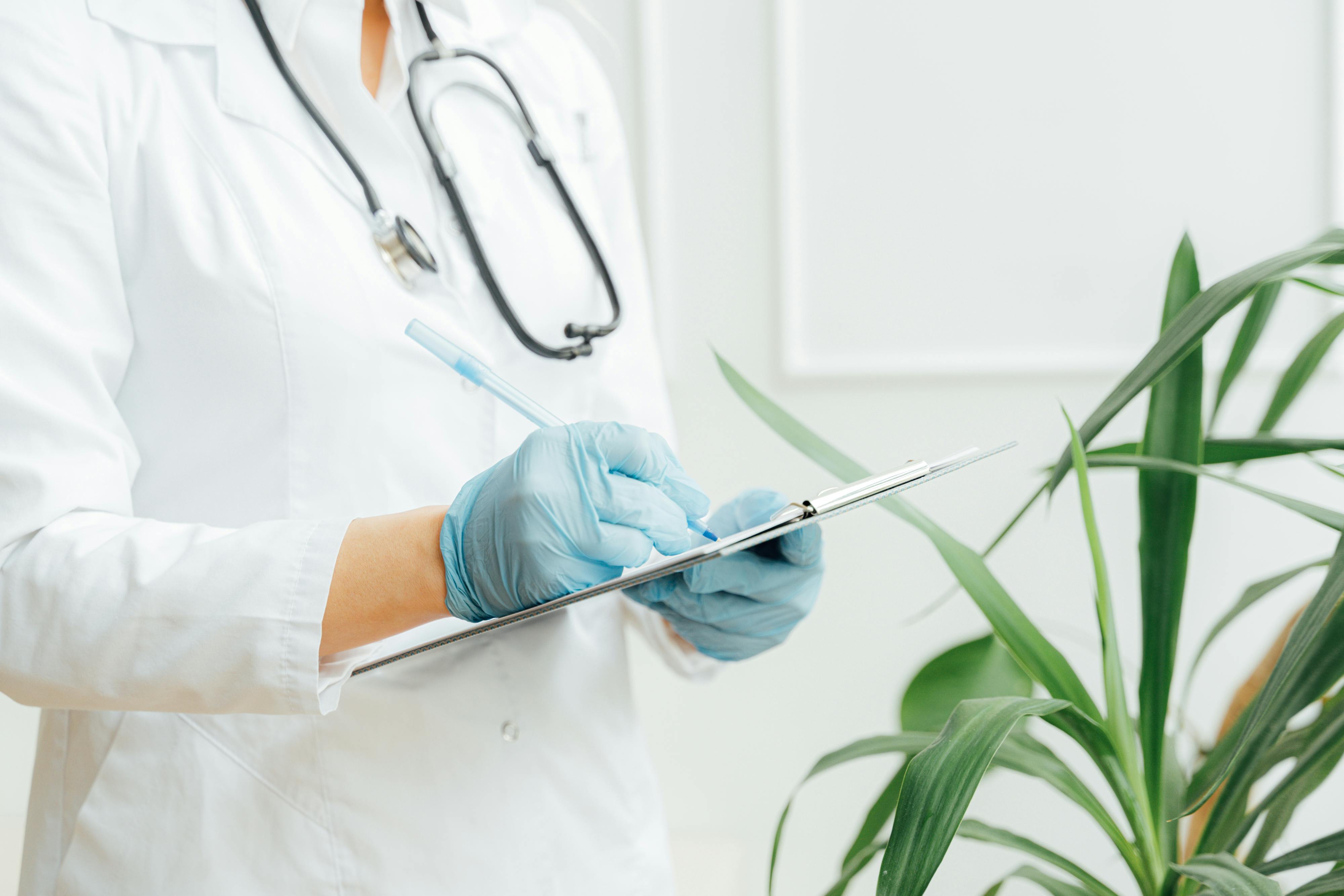 Uma pessoa médica com uma prancheta na mão, vestida com jaleco, luvas azuis e com um estetoscópio no pescoço, representando a atenção que deve ser dada para a medicina e os programas d esaúde dentro das empresas.
